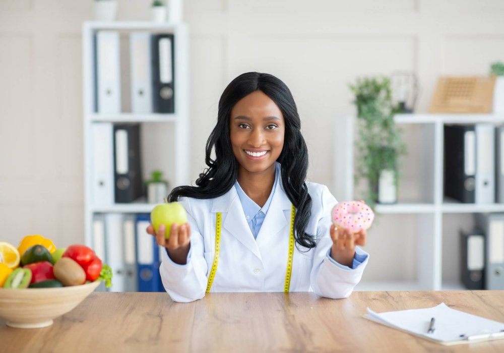 Pretty black dietary doctor holding donut and apple at her office, offering choice between fresh and